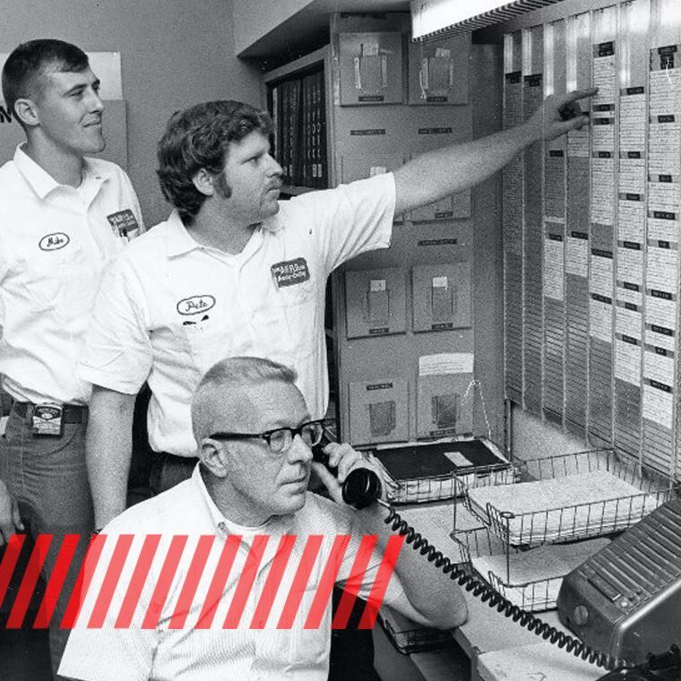 Three men in a vintage office: one on the phone, two examining a large information board.